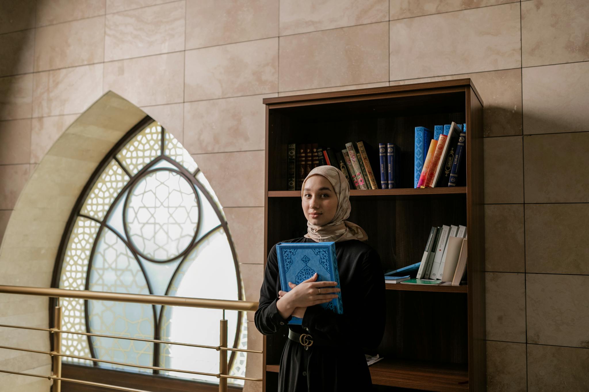 Young woman in hijab holding a Quran in a beautifully lit library with arched windows.